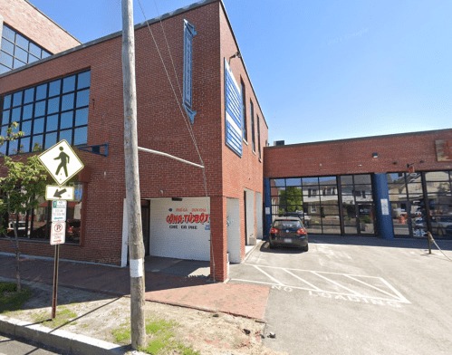 A brick building with large windows, featuring a parking area and a "No Parking" sign.