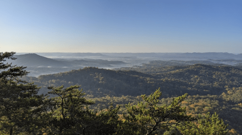 A panoramic view of misty mountains and valleys under a clear blue sky, with lush greenery in the foreground.