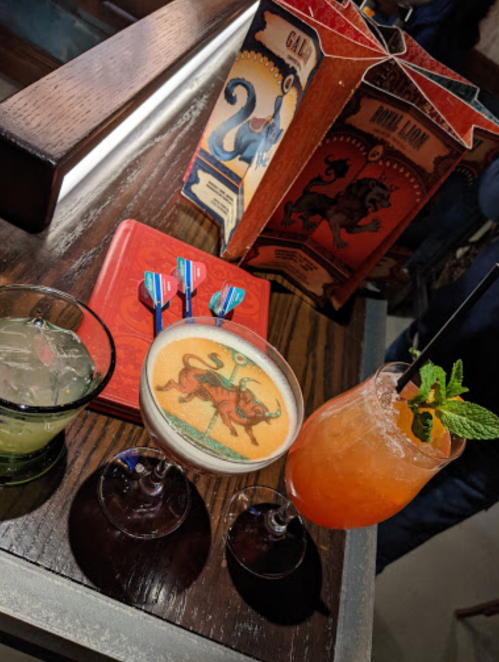 Three colorful cocktails on a wooden table, with a menu in the background featuring a lion and octopus design.