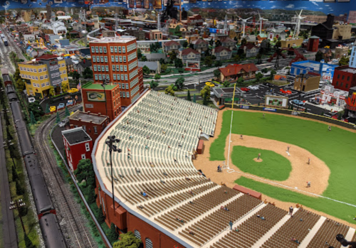 A detailed miniature model of a baseball stadium with surrounding buildings and a train track in the background.