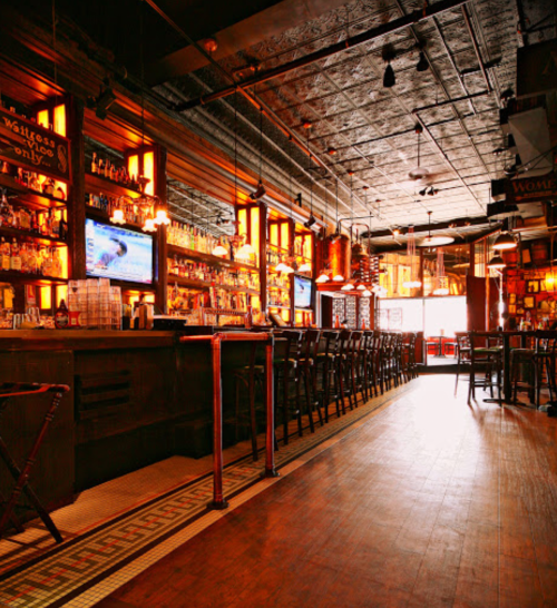A cozy bar interior with wooden furnishings, shelves of liquor, and warm lighting, creating an inviting atmosphere.