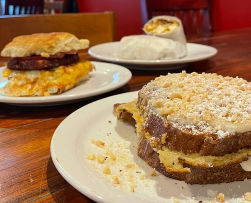 A plate with three breakfast items: a sandwich, a wrap, and a slice of French toast topped with powdered sugar and nuts.