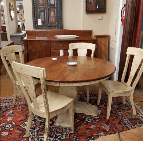A round wooden dining table with four distressed white chairs, set on a patterned rug in a cozy interior.