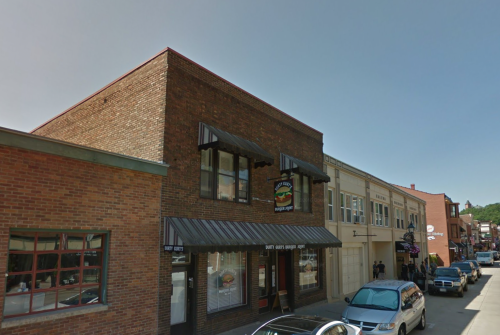 A brick building with a sign for a burger shop, surrounded by other shops and parked cars on a sunny day.