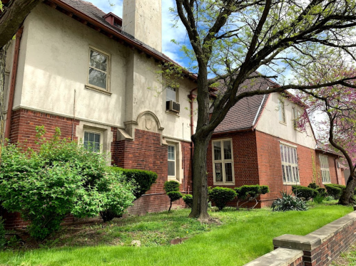 A brick building with a white facade, surrounded by green grass and trees, under a partly cloudy sky.