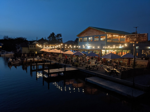A waterfront restaurant at dusk, with outdoor seating and string lights illuminating the patio.