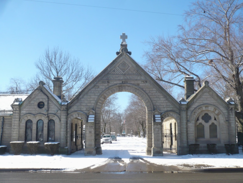Stone archway entrance to a cemetery, flanked by trees and covered in snow under a clear blue sky.
