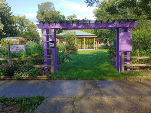 A purple garden archway leads to a lush green space with a gazebo and flower beds in the background.