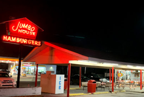 A brightly lit fast-food restaurant at night, featuring a red neon sign that reads "Jumbo House Hamburgers."