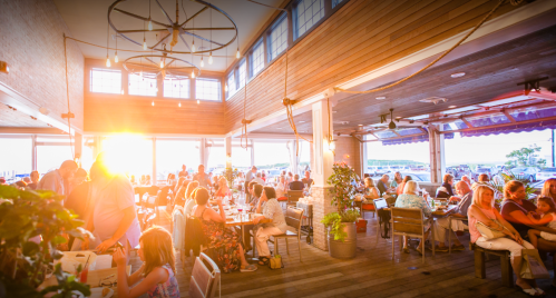 A lively restaurant interior filled with diners, bathed in warm sunset light, creating a vibrant atmosphere.