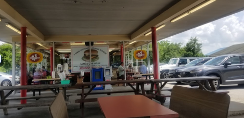 Outdoor dining area with picnic tables, a food stand, and parked cars under a bright sky.