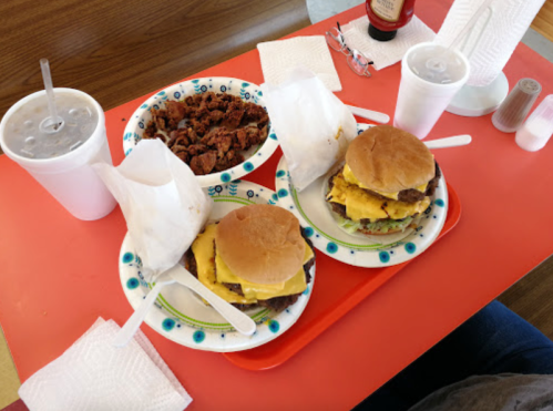 Two cheeseburgers with lettuce and pickles on plates, accompanied by a bowl of snacks and drinks on a red table.