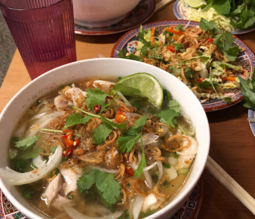A bowl of pho with herbs, lime, and chili, alongside a plate of salad and a pink drink in a glass.