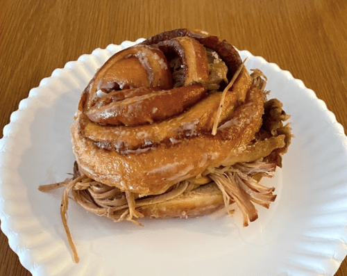 A cinnamon roll topped with pulled pork, served on a white paper plate.