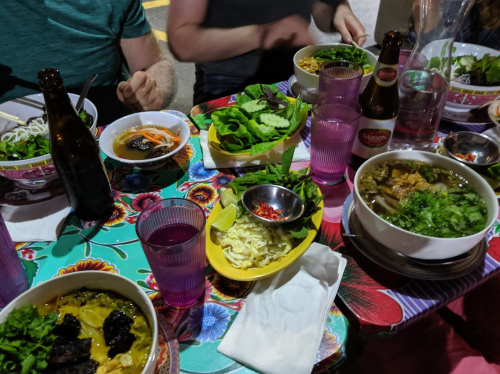 A vibrant table filled with various bowls of noodles, fresh herbs, and drinks, set in a lively dining atmosphere.