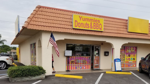 Exterior of "Yummies Donuts & BBQ" shop with a sign, American flag, and colorful window displays.