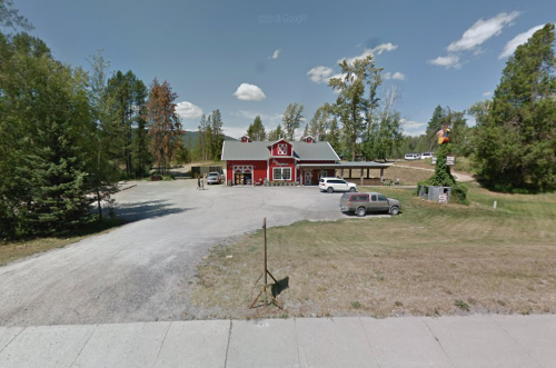 A red building with a large porch, surrounded by trees and parked cars, set in a rural area under a blue sky.