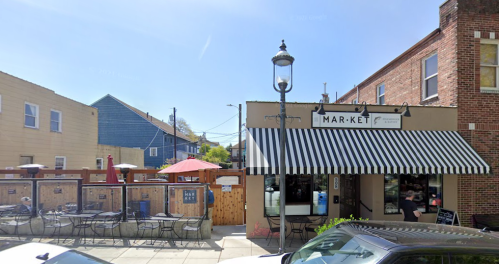 A street view of a market with outdoor seating, striped awning, and nearby buildings under a clear blue sky.