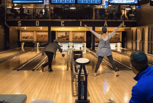 Two people bowling in a lively alley, one celebrating a strike while the other prepares to roll. Bright lights and screens in the background.