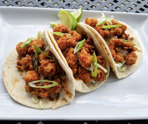 Three crispy fried chicken tacos topped with green onions and served with lime wedges on a white plate.