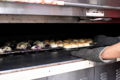 A person wearing oven mitts pulls a tray of baked goods from an oven, showcasing muffins and pastries.