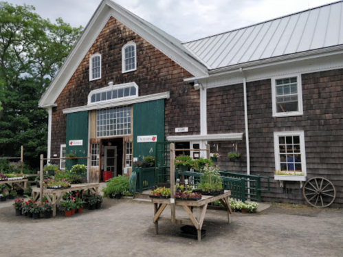 A rustic barn with green doors, surrounded by flower pots and plants for sale, under a cloudy sky.
