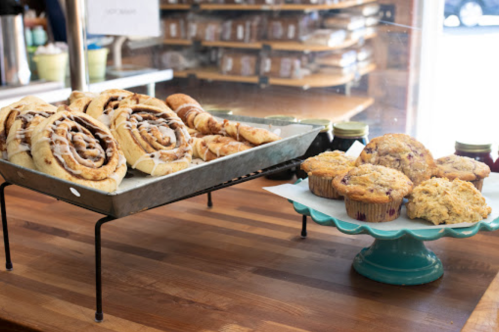 A display of baked goods: cinnamon rolls on a tray and muffins on a stand, with jars of jam in the background.