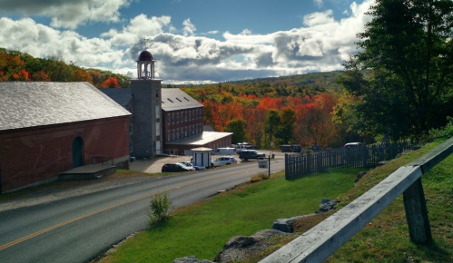 A scenic view of a red building beside a road, surrounded by vibrant autumn foliage and a cloudy sky.