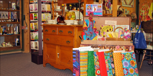 A cozy shop interior featuring colorful fabrics, a wooden dresser, and various crafts displayed on shelves.