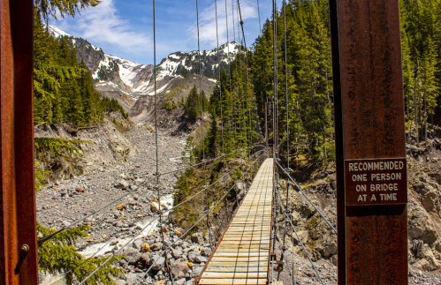A narrow suspension bridge spans a rocky riverbed, surrounded by lush trees and snow-capped mountains in the background.