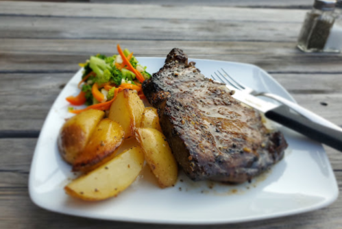 A plate of grilled steak with roasted potatoes and a side of mixed vegetables on a wooden table.