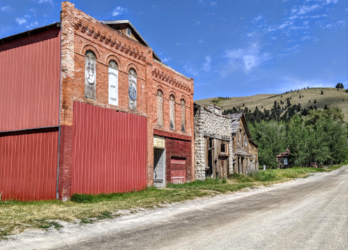 Abandoned buildings along a dirt road, with a blue sky and green hills in the background.
