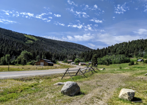 A scenic view of a rural road with wooden fences, surrounded by green hills and trees under a blue sky.