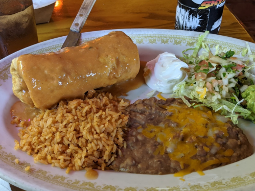 A plate featuring a burrito with sauce, rice, refried beans, salad, and sour cream.