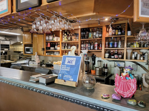 A cozy bar area with shelves of drinks, glasses hanging, and a sign indicating the business is open.
