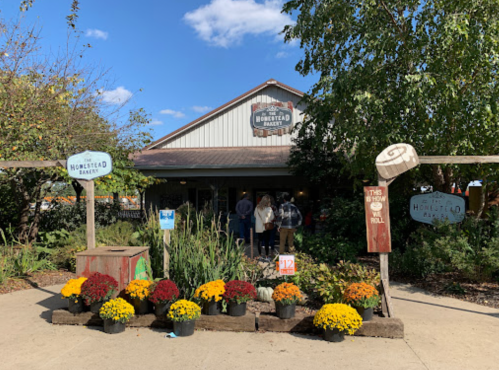 A rustic bakery with a sign, surrounded by colorful flowers and greenery, under a blue sky with a few clouds.