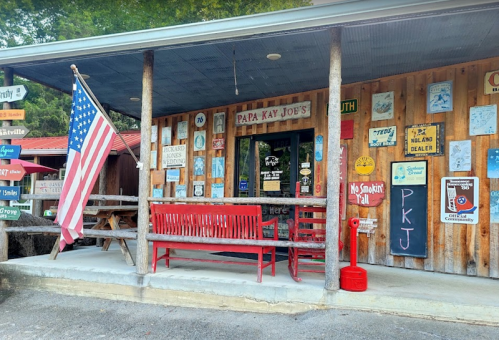 A rustic storefront with a red bench, American flag, and various signs on a wooden exterior.