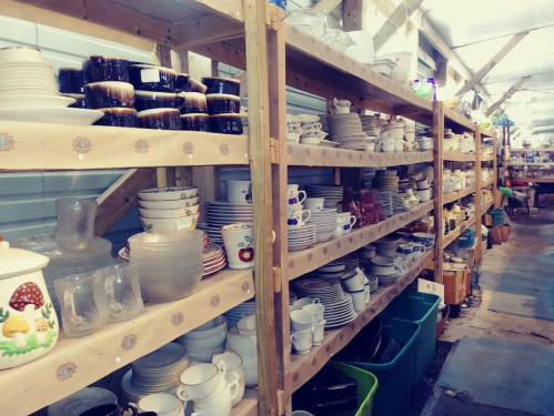 Aisle of shelves filled with various dishes, cups, and glassware in a market or thrift store.