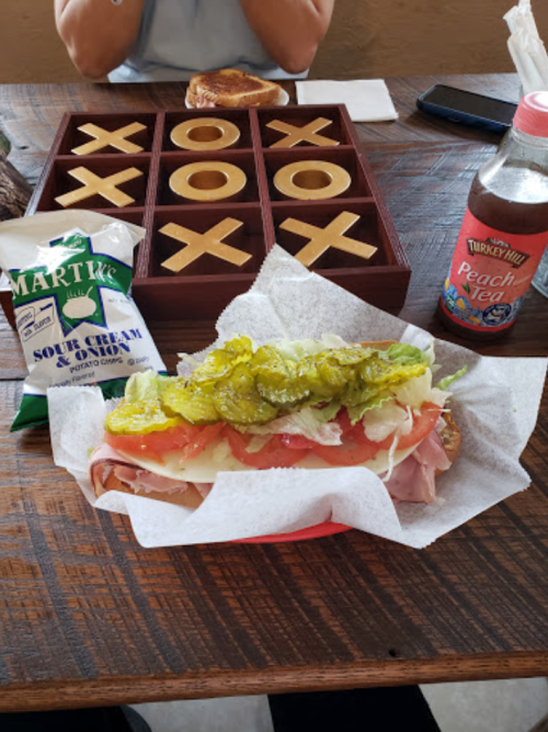 A sandwich with lettuce, tomato, and pickles on a table with a game of tic-tac-toe and a drink.