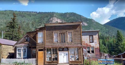 A rustic wooden building with a porch, surrounded by trees and mountains under a blue sky with clouds.
