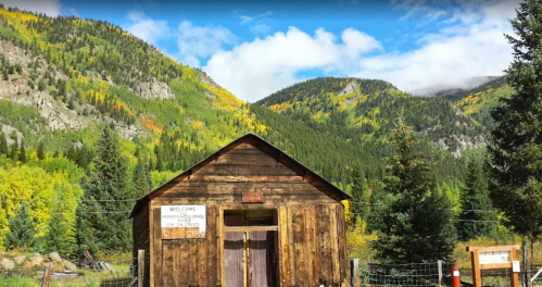 A rustic wooden cabin surrounded by vibrant autumn foliage and mountains under a partly cloudy sky.