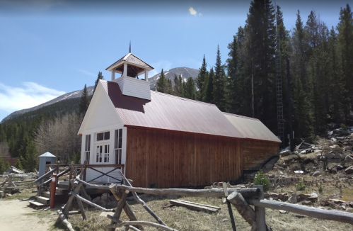 A rustic wooden building with a red roof, surrounded by trees and mountains, featuring a small bell tower.