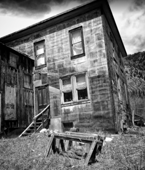 A tilted, weathered two-story house with boarded windows, set against a mountainous backdrop in black and white.