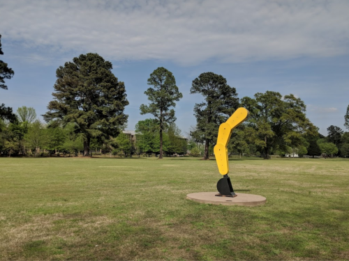 A bright yellow abstract sculpture stands on a circular base in a grassy park, surrounded by trees and blue sky.