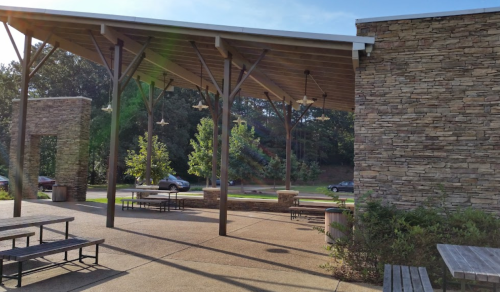 Outdoor seating area with stone walls, a wooden roof, and trees in the background on a sunny day.
