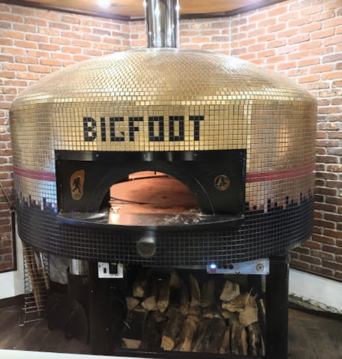 A large, gold-tiled pizza oven with "BIGFOOT" written on it, set against a brick wall. Logs are stacked below.