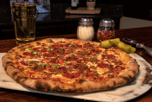 A freshly baked pizza topped with pepperoni, sausage, green peppers, and served with beer, condiments, and pickles.