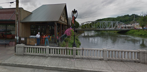 A riverside view of a restaurant with colorful seating, a bridge in the background, and lush greenery along the water.