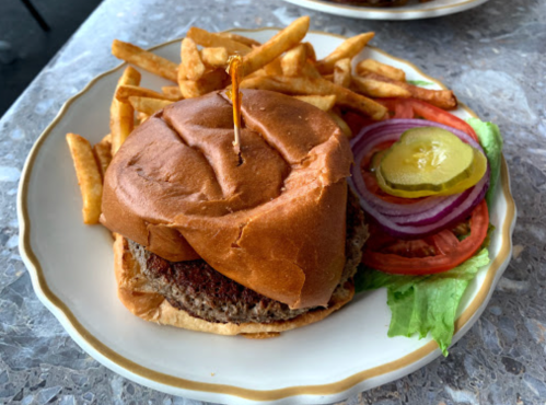 A plate with a burger topped with a toothpick, served with fries, lettuce, tomato, onion, and a pickle slice.