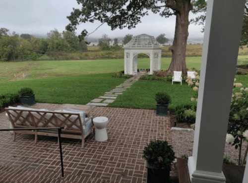 A serene outdoor scene featuring a gazebo, stone path, and seating area on a grassy lawn surrounded by trees.